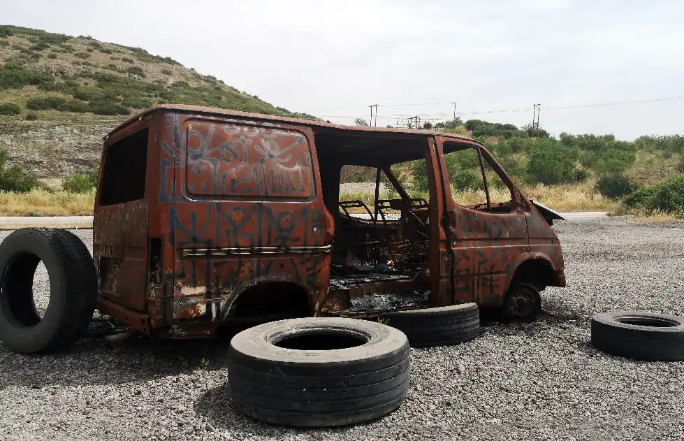A heavily rusted and burnt out van covered in graffiti abandoned on a gravel roadside with old tyres scattered around it.