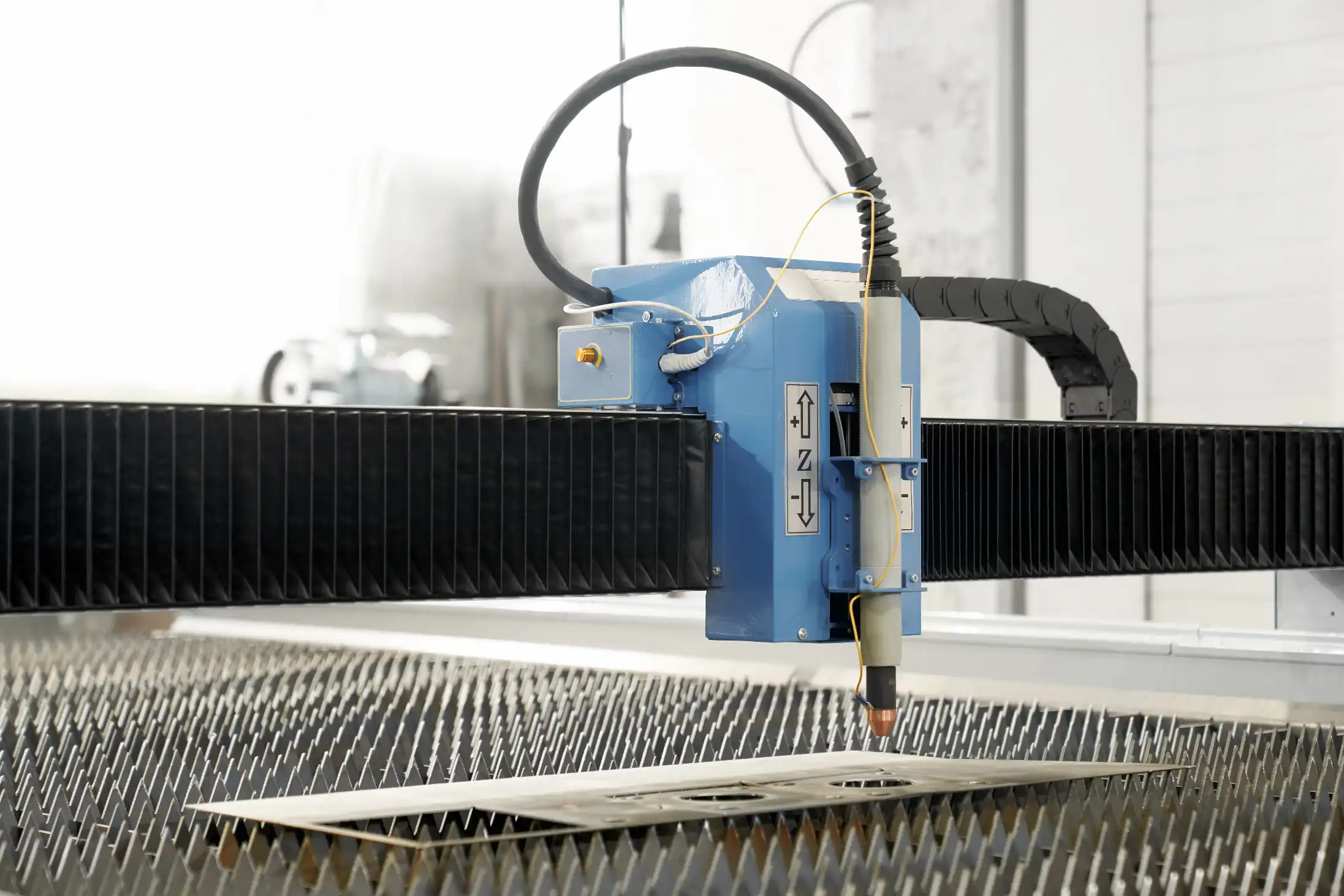 A blue CNC plasma laser cutting machine head moving over a metal sheet on a factory cutting table showing the precision cutting process.