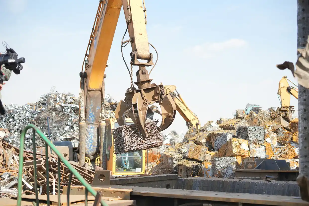 A yellow Hyundai grapple crane lifting twisted metal rods and wire at a large industrial scrap metal recycling facility.