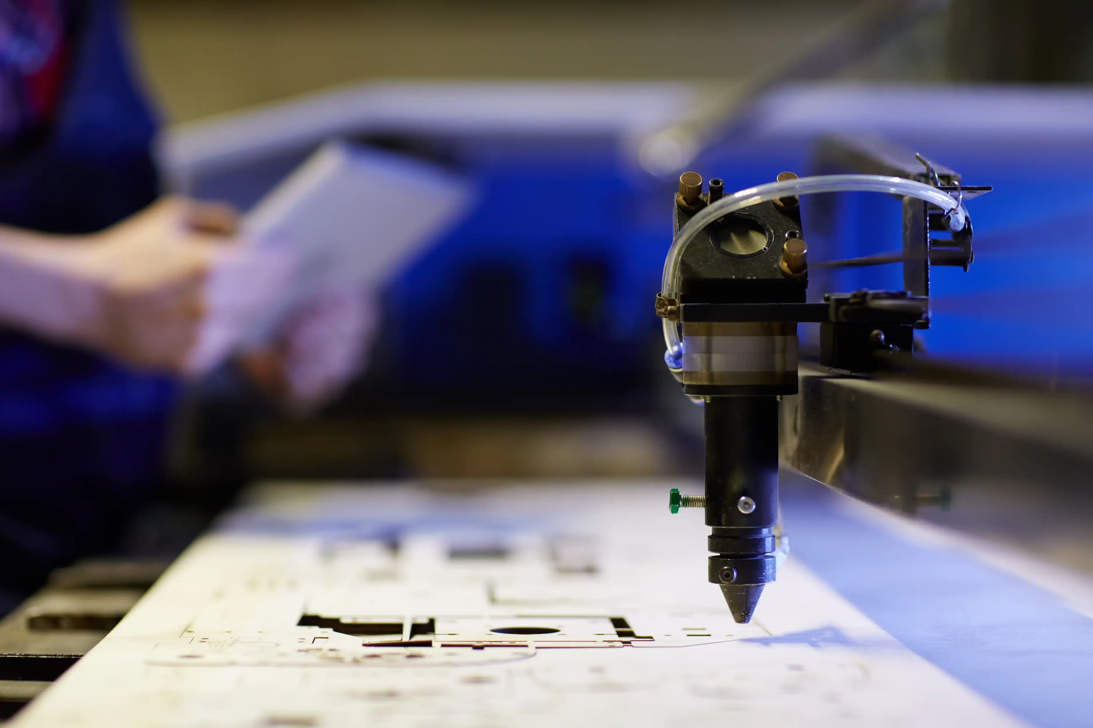 A close-up of an industrial laser cutting head cutting precise shapes into a metal sheet while a worker holds documents in the background.