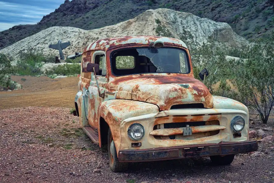 A heavily rusted and deteriorated old truck with broken windows abandoned in a rocky desert landscape showing a classic end-of-life vehicle.