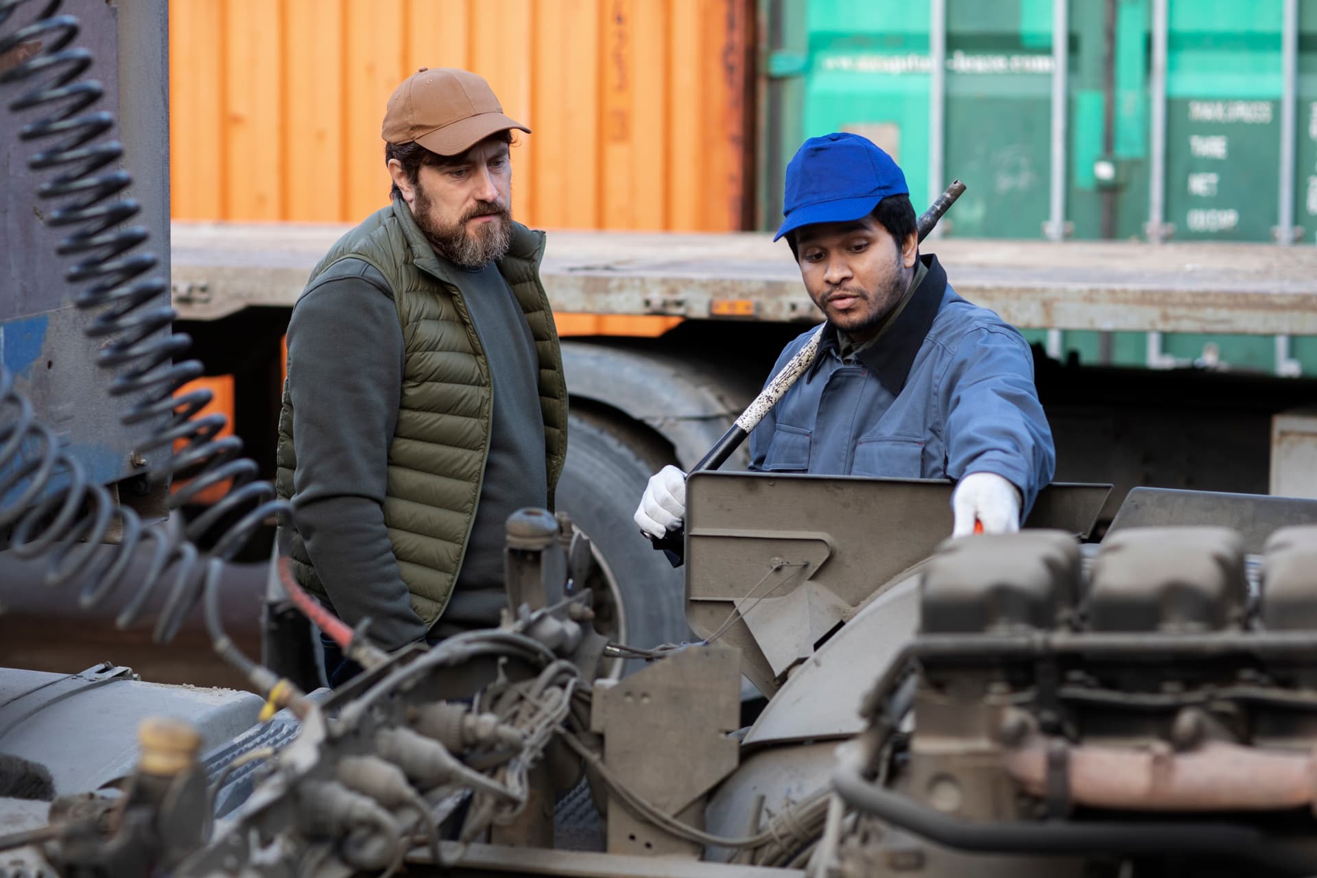 Two workers inspecting scrap metal parts at an outdoor yard next to a truck, deciding when to sell.