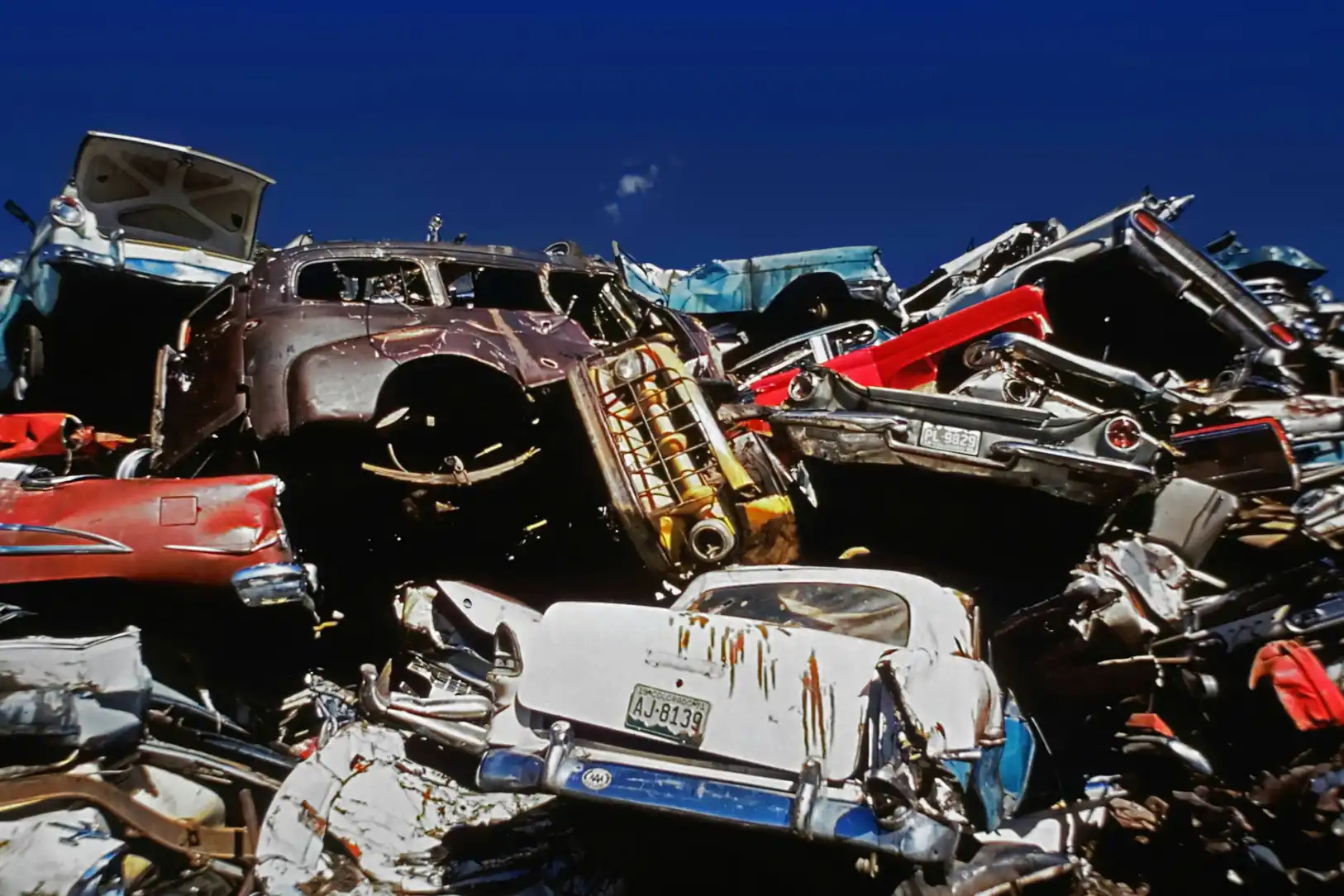 A pile of crushed and rusted vintage cars stacked on top of each other at a scrapyard under a clear blue sky.